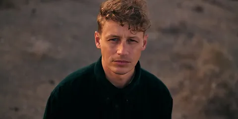 Junger Mann mit blonden locken schaut den Btrachter direkt an.