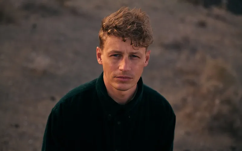 Junger Mann mit blonden locken schaut den Btrachter direkt an.