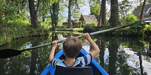 Ein Junge paddelt mit einem blauen Kajak auf einem Spreekanal in Burg im Spreewald. Die Umgebung ist von satten Grüntönen geprägt. Im Hintergrund ist ein traditionelles Spreewaldhaus zu sehen.