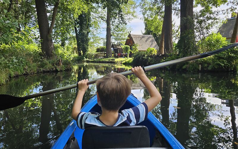Ein Junge paddelt mit einem blauen Kajak auf einem Spreekanal in Burg im Spreewald. Die Umgebung ist von satten Grüntönen geprägt. Im Hintergrund ist ein traditionelles Spreewaldhaus zu sehen.