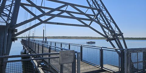 Zu sehen ist ein imposanter Steg, der Teil eines großen Baggergeräts war. Im Hintergrund fährt ein Schiff über den blauen See. Darüber erstreckt sich ein blauer Himmel.
