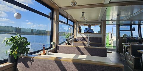 Zu sehen ist das Unterdeck des Schiffes mit hellen Tischen und braunen Bänken. Durch die Fenster bieten sich Ausblicke auf den blauen Himmel und das blaue Wasser des Bärwalder Sees.