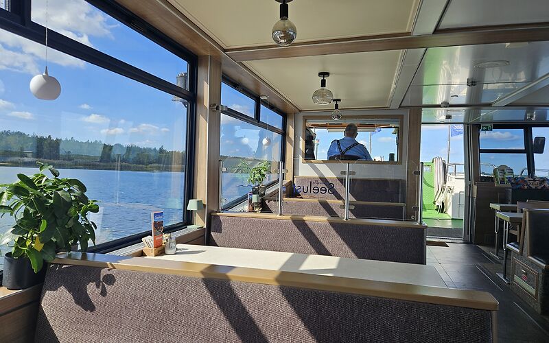 Zu sehen ist das Unterdeck des Schiffes mit hellen Tischen und braunen Bänken. Durch die Fenster bieten sich Ausblicke auf den blauen Himmel und das blaue Wasser des Bärwalder Sees.