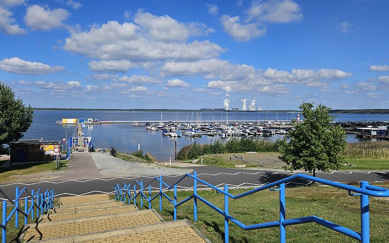 Blick auf den Hafen am Bärwalder See mit zahlreichen Booten. Im Vordergrund führt eine breite Treppe mit blauem Geländer vom Parkplatz hinunter zum Hafen.
