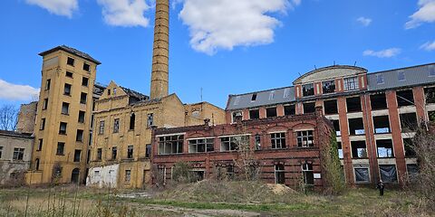 Das Foto zeigt die historische Industrieanlage in Forst (Lausitz). Zu sehen sind alte Fabrikgebäude aus rotem und gelbem Backstein, ein hoher Schornstein sowie teilweise verfallene Hallen mit großen Fenstern. Vor den Gebäuden befindet sich eine bewachsene Freifläche.
