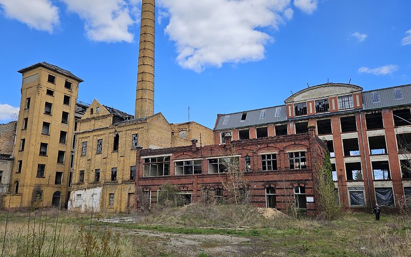 Das Foto zeigt die historische Industrieanlage in Forst (Lausitz). Zu sehen sind alte Fabrikgebäude aus rotem und gelbem Backstein, ein hoher Schornstein sowie teilweise verfallene Hallen mit großen Fenstern. Vor den Gebäuden befindet sich eine bewachsene Freifläche.