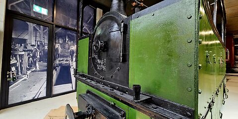 Zu sehen ist die historische Lokomotive „Schwarze Jule“ aus dem Jahr 1892 in den Farben Grün und Schwarz. Sie ist zusätzlich auf einer großen schwarz-weißen Fotowand abgebildet, auf der Mitarbeitende bei der Durchsicht der Lokomotive zu sehen sind.