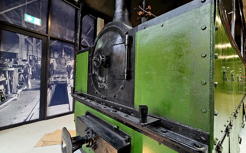 Zu sehen ist die historische Lokomotive „Schwarze Jule“ aus dem Jahr 1892 in den Farben Grün und Schwarz. Sie ist zusätzlich auf einer großen schwarz-weißen Fotowand abgebildet, auf der Mitarbeitende bei der Durchsicht der Lokomotive zu sehen sind.