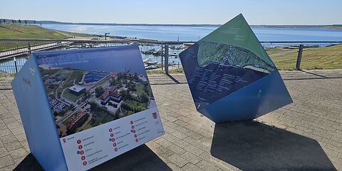 Auf dem Bild sind zwei große Informationswürfel zum Großräschener See zu sehen. Auf den Seiten der Würfel befinden sich Fotos und Informationen über den See. Im Hintergrund ist bei sonnigem Wetter der blau schimmernde See unter einem klaren, blauen Himmel zu sehen.