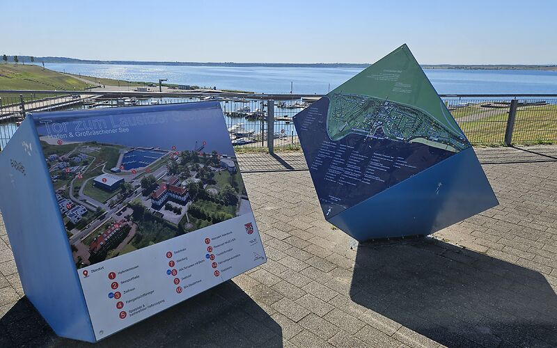 Auf dem Bild sind zwei große Informationswürfel zum Großräschener See zu sehen. Auf den Seiten der Würfel befinden sich Fotos und Informationen über den See. Im Hintergrund ist bei sonnigem Wetter der blau schimmernde See unter einem klaren, blauen Himmel zu sehen.