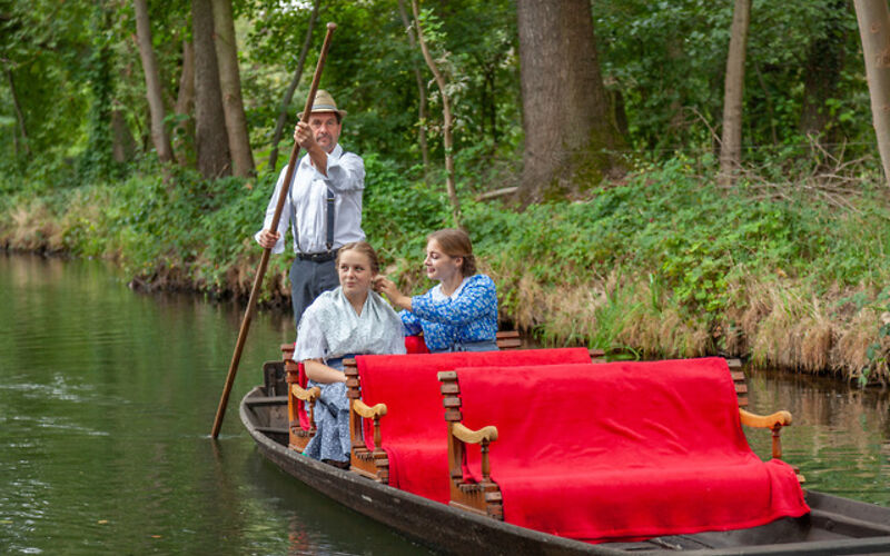 Zwei Frauen in blauen regionalen Trachten unternehmen gemeinsam mit einem Kahnfährmann eine Fahrt in einem traditionellen Holzkahn. Die Sitzbänke sind mit roten Decken geschmückt. Im Hintergrund sind grüne Bäume zu sehen.