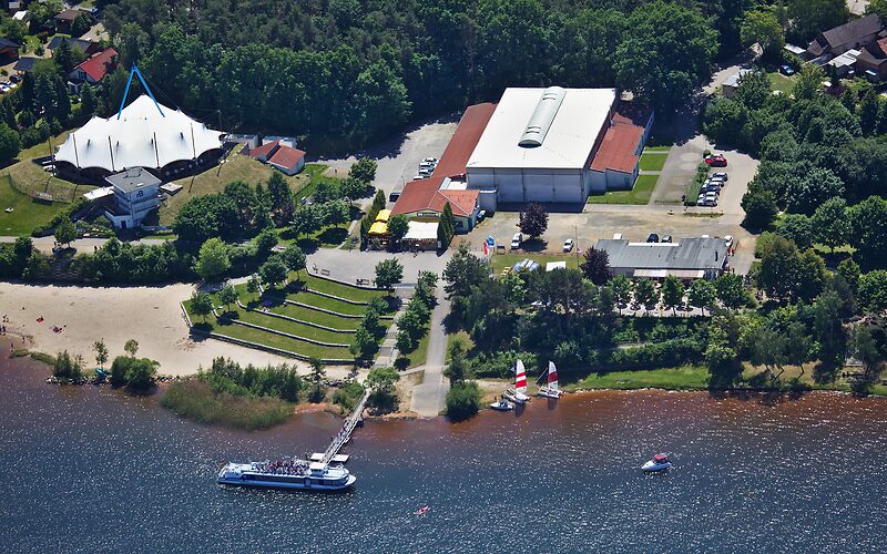 Luftaufnahme des Hafens am Senftenberger See in Großkoschen. Aus der Vogelperspektive sind das große Veranstaltungszelt, weitere Gebäude des Hafengeländes, der Strand und der See zu sehen. Im Hafen liegt das Schiff „St. Barbara“. 