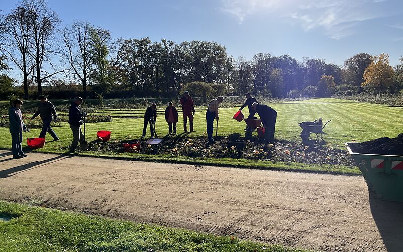 Mehrer Menschen stehen bei einem Rosenbeet an einem Sandweg. Sie haben Gartenwerkzeuge in den Händen. Schubkaaren und Eimer stehen dabenen. Die Sonne scheint hell und im Hintergrund sind viele Bäume zu sehen.  