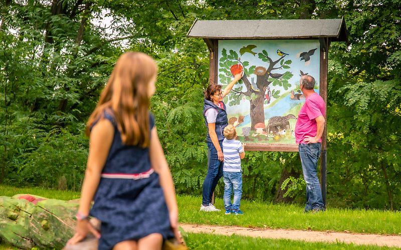 Mädchen sitz im Vordergrund und schaut nach hinten, im Hitergrund steht Familie schut interessiert auf eine Schautafel