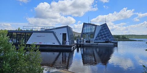 Auf dem Foto sind zwei moderne Gebäude auf dem Bergheider See zu sehen: ein Aquaforum und ein schwimmendes Haus mit Solaranlage.