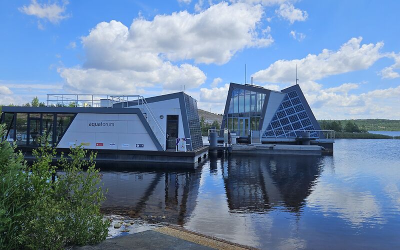 Auf dem Foto sind zwei moderne Gebäude auf dem Bergheider See zu sehen: ein Aquaforum und ein schwimmendes Haus mit Solaranlage.