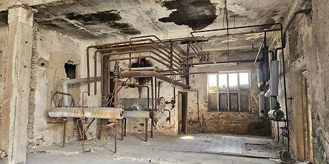 Blick in einen verlassenen Innenraum einer Industrieanlage mit verwitterten Wänden, einer alten Decke, einem alten Fenster und sichtbaren Rohrleitungen der ehemaligen Anlage.