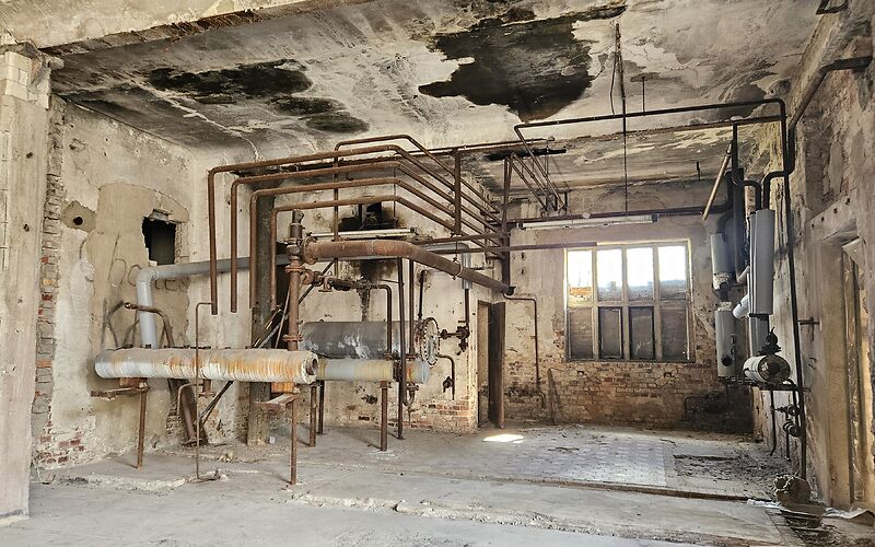 Blick in einen verlassenen Innenraum einer Industrieanlage mit verwitterten Wänden, einer alten Decke, einem alten Fenster und sichtbaren Rohrleitungen der ehemaligen Anlage.