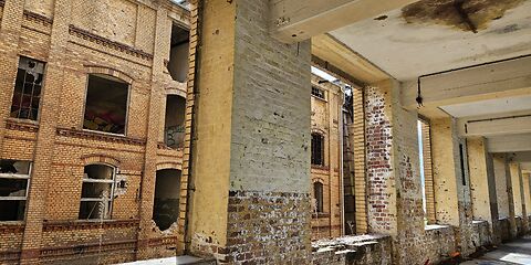 Blick auf den Innenbereich einer verlassenen Industrieanlage. Sichtbar sind alte Backsteinmauern, große Fensteröffnungen und massive Pfeiler mit Spuren des Verfalls.