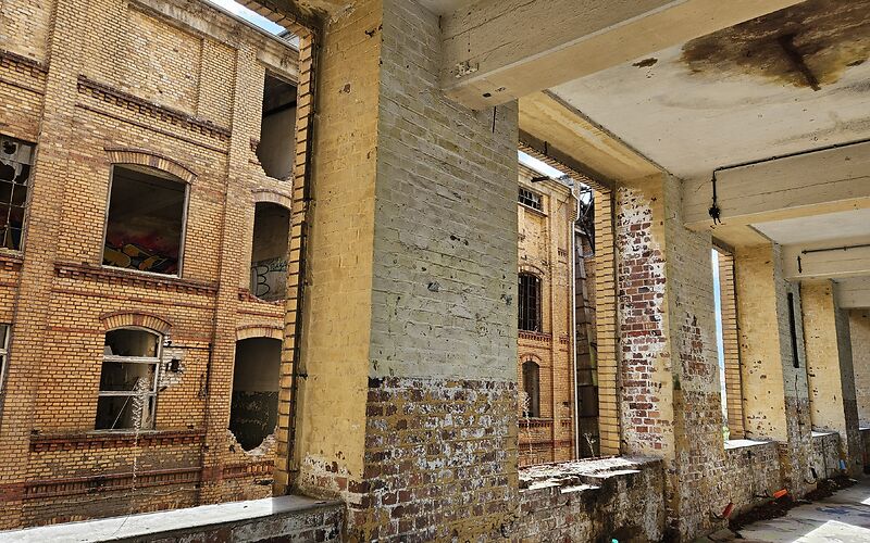 Blick auf den Innenbereich einer verlassenen Industrieanlage. Sichtbar sind alte Backsteinmauern, große Fensteröffnungen und massive Pfeiler mit Spuren des Verfalls.