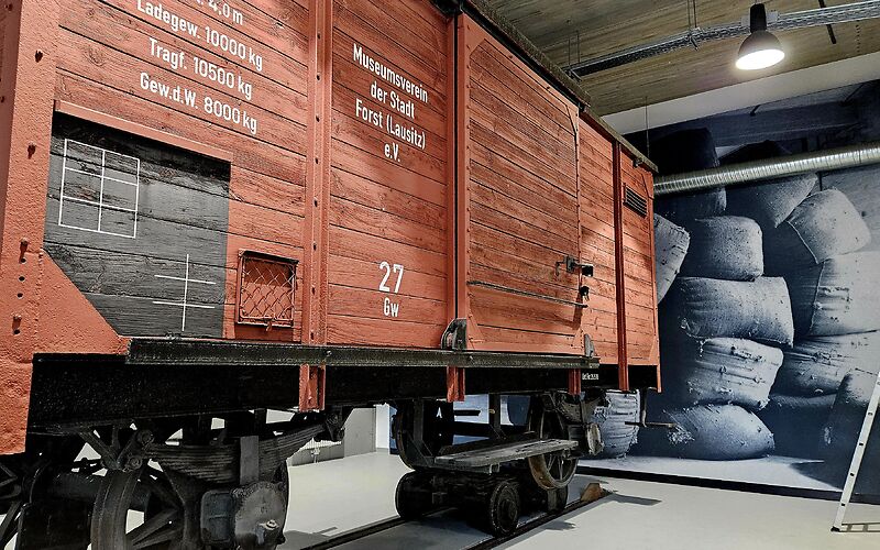 Im Vordergrund steht ein rotbrauner Kohlenwagen, der auf Rollböcken eingesetzt ist. Auf dem Wagen sind die Aufschriften „Ladegewicht 10.000 kg“, „Tragfähigkeit 10.500 kg“ sowie „Museumsverein der Stadt Forst (Lausitz)“ zu lesen. Im Hintergrund ist eine große schwarz-weiße Fotowand mit voll gefüllten Säcken zu sehen.