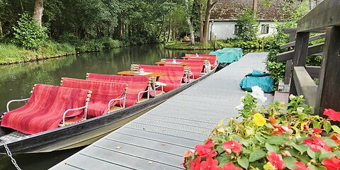Ein Kahn mit Tischen und Bänken, die mit roten Decken bedeckt sind, liegt auf einem Kanal im Spreewald in Burg. Die grüne, idyllische Umgebung prägt die Szenerie.