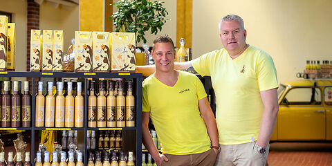 Die Besitzer der Manufaktur scharfesGELB stehen im Verkaufsraum neben bunten Eierlikörflaschen. Sie tragen gelbe T-Shirts, die Flaschen sind auf Regalen aufgereiht.
