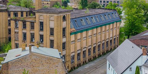 Auf dem Foto ist die frühere dreietagige Fabrik C. H. Pürschel zu sehen. Das Gebäude besteht aus hellen Ziegeln. Vor dem Gebäude verlaufen Schienen.