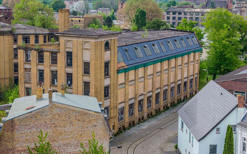 Auf dem Foto ist die frühere dreietagige Fabrik C. H. Pürschel zu sehen. Das Gebäude besteht aus hellen Ziegeln. Vor dem Gebäude verlaufen Schienen.