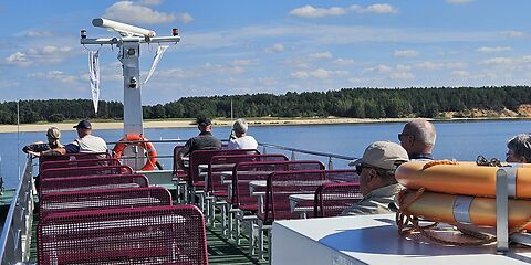 Sonnige Schifffahrt auf dem Oberdeck des Ausflugsschiffes. Einige Touristen sitzen an Tischen und genießen die Fahrt. Der blaue Himmel mit weißen Wolken, das blaue Wasser und der Strand im Hintergrund sorgen für eine wunderschöne Atmosphäre.