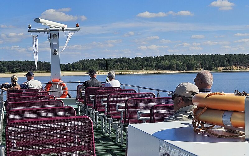 Sonnige Schifffahrt auf dem Oberdeck des Ausflugsschiffes. Einige Touristen sitzen an Tischen und genießen die Fahrt. Der blaue Himmel mit weißen Wolken, das blaue Wasser und der Strand im Hintergrund sorgen für eine wunderschöne Atmosphäre.