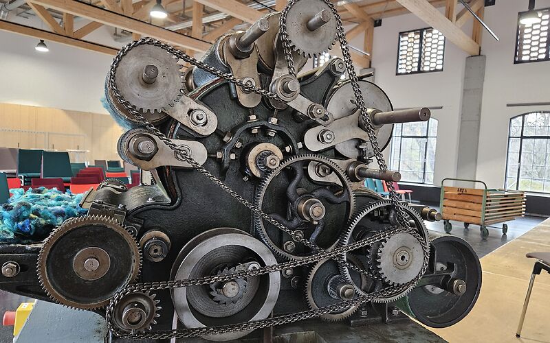 Zu sehen ist ein großes Maschinenteil einer historischen Textilmaschine in einem modernen Veranstaltungsraum mit großen Fenstern. An dem Maschinenteil sind Wollfasern in verschiedenen Blautönen angebracht.