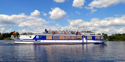 Ein weiß-blaues Schiff fährt über den Senftenberger See. Am Himmel sind weiße Wolken zu sehen, die sich vom blauen Himmel abheben. Die Sonne lässt das Wasser hell glänzen und sorgt für eine freundliche, sommerliche Atmosphäre.