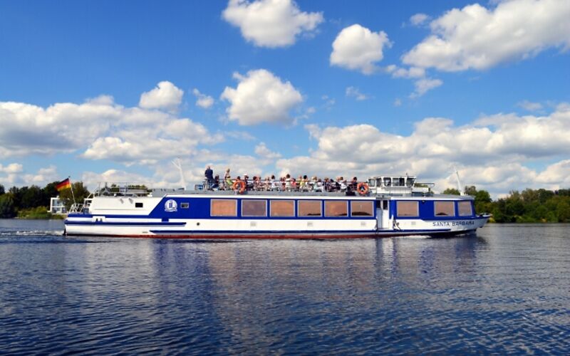 Ein weiß-blaues Schiff fährt über den Senftenberger See. Am Himmel sind weiße Wolken zu sehen, die sich vom blauen Himmel abheben. Die Sonne lässt das Wasser hell glänzen und sorgt für eine freundliche, sommerliche Atmosphäre.