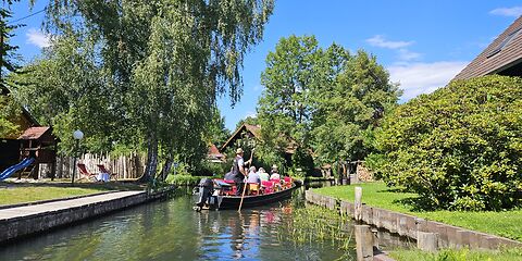 Kahn mit mehreren Personen auf einem Spreewaldkanal in Lehde, dahinter von Bäumen verdeckte Spreewaldhäuser und blauer Himmel.