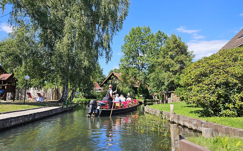 Kahn mit mehreren Personen auf einem Spreewaldkanal in Lehde, dahinter von Bäumen verdeckte Spreewaldhäuser und blauer Himmel.
