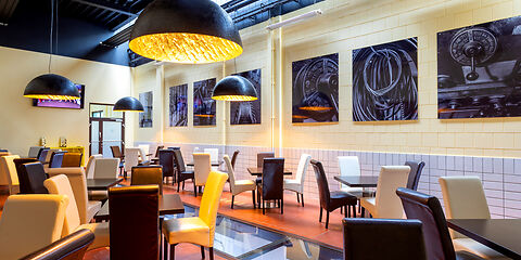 Ein Caféraum mit vielen Tischen und Stühlen. Die Stühle sind bunt, die Farben im Raum sind Gelb, Braun und Schwarz.