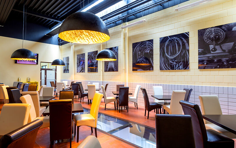 Ein Caféraum mit vielen Tischen und Stühlen. Die Stühle sind bunt, die Farben im Raum sind Gelb, Braun und Schwarz.