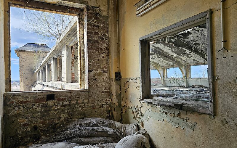 Blick in einen verlassenen Innenraum einer Industrieanlage mit verwitterten Wänden, alten Fensteröffnungen und sichtbaren Spuren des Verfalls. Durch die Fenster sind weitere Gebäudeteile der Fabrikanlage zu sehen.