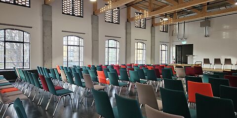 Zu sehen ist der moderne, helle Veranstaltungsraum des Forster Museums für Textil- und Industriegeschichte Lausitz. Der Raum hat große Fenster und ist mit vielen bunten Stühlen ausgestattet, darunter rote, grüne, schwarze und braune Sitzplätze.