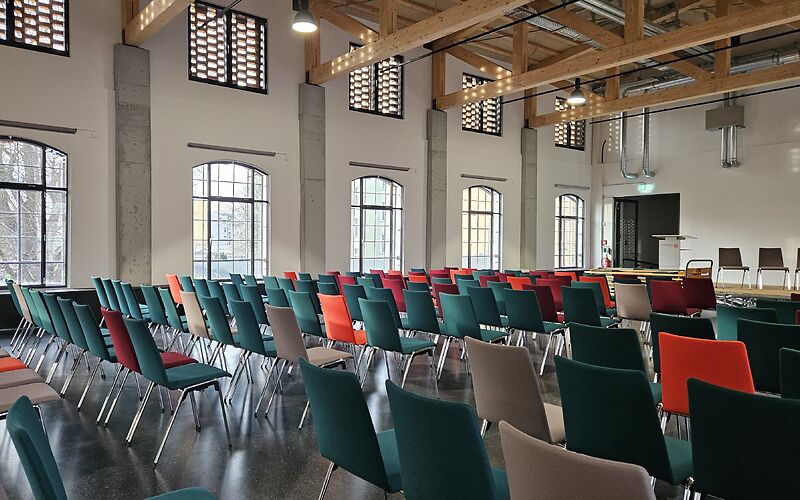Zu sehen ist der moderne, helle Veranstaltungsraum des Forster Museums für Textil- und Industriegeschichte Lausitz. Der Raum hat große Fenster und ist mit vielen bunten Stühlen ausgestattet, darunter rote, grüne, schwarze und braune Sitzplätze.