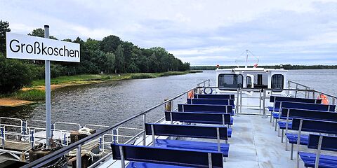 Das Fahrgastschiff „Santa Barbara“ liegt im Hafen von Großkoschen am Senftenberger See. Auf dem Oberdeck befinden sich blaue Sitzbänke. Ein großes Schild mit dem Namen des Ortes weist auf den Hafenstandort hin.