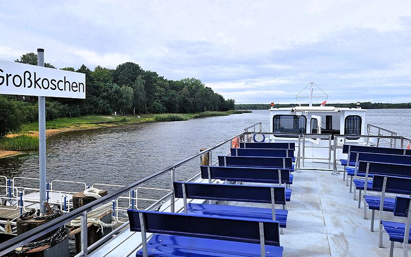 Das Fahrgastschiff „Santa Barbara“ liegt im Hafen von Großkoschen am Senftenberger See. Auf dem Oberdeck befinden sich blaue Sitzbänke. Ein großes Schild mit dem Namen des Ortes weist auf den Hafenstandort hin.