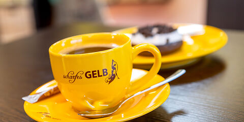 Gelbe Kaffeetasse mit dem Logo "scharfesGELB". Daneben liegt ein gelber Teller mit einem Keks.