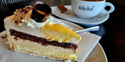 Eine dreifarbige Torte mit Eierlikör wird zusammen mit einer Tasse Kaffee serviert. Der Eierlikör wird in einem kleinen Becher aus Zartbitterschokolade gereicht. Die Tasse trägt den Schriftzug „Confiserie Felicitas“.