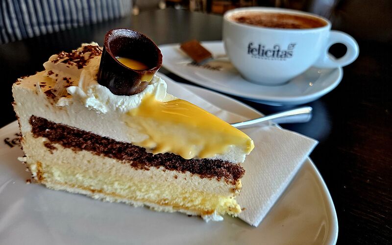 Eine dreifarbige Torte mit Eierlikör wird zusammen mit einer Tasse Kaffee serviert. Der Eierlikör wird in einem kleinen Becher aus Zartbitterschokolade gereicht. Die Tasse trägt den Schriftzug „Confiserie Felicitas“.