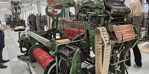 Das Foto zeigt eine Reihe von Textilmaschinen, die überwiegend in Grün sowie Schwarz-Silber gehalten sind. Im Vordergrund ist eine Maschine zu sehen, auf der sich rote Wollfäden auf einer großen Spule befinden.