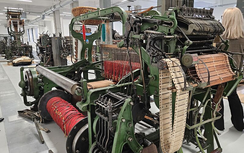 Das Foto zeigt eine Reihe von Textilmaschinen, die überwiegend in Grün sowie Schwarz-Silber gehalten sind. Im Vordergrund ist eine Maschine zu sehen, auf der sich rote Wollfäden auf einer großen Spule befinden.