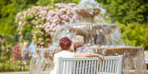 Auf dem Foto sitzt ein Paar auf einer weißen Bank vor dem Kaskadenbrunnen im Rosengarten. Umgeben von blühenden Rosen genießt es die sonnige Atmosphäre des Gartens.