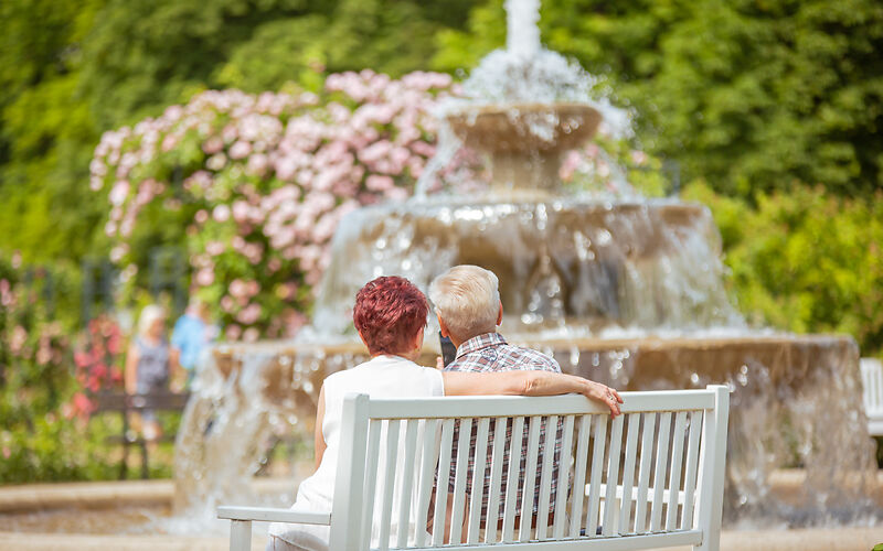 Auf dem Foto sitzt ein Paar auf einer weißen Bank vor dem Kaskadenbrunnen im Rosengarten. Umgeben von blühenden Rosen genießt es die sonnige Atmosphäre des Gartens.